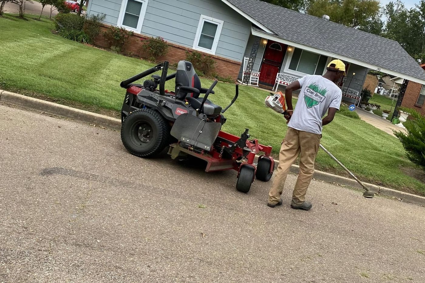 A Clark Bros crew member in a company shirt string-trimming a front lawn beside a zero-turn mower